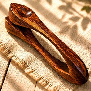 May include: Wooden serving tongs with a dark brown finish. The tongs are made from a single piece of wood, featuring a curved handle and a scoop-shaped head. They rest on a tan, textured surface, likely linen or a similar fabric.