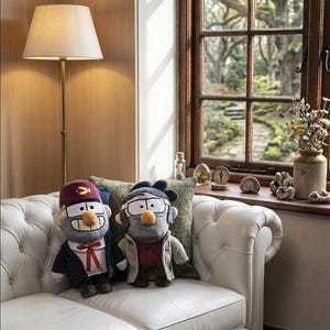 May include: Two plush dolls representing characters from Gravity Falls. One doll wears a dark suit, white shirt, red bolo tie, and a maroon fez with a gold symbol. The other doll wears a beige coat, dark shirt, and glasses with thick frames.