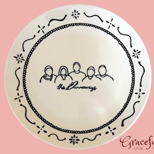 May include: A white ceramic plate with a black outline design. The plate features a black border with decorative elements and the text "the Runaways" with a line drawing of five people. The background is pink.