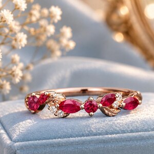 Puede incluir: Un anillo de oro rosa con una serie de rubíes rojos en talla marquesa y redonda, intercalados con pequeñas piedras transparentes y detalles metálicos en forma de hoja. El anillo descansa sobre una caja de terciopelo azul claro con el texto Bijouraus.