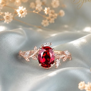 Könnte beinhalten: Ein Ring aus Roségold mit einem zentralen, oval geschliffenen, tiefroten Edelstein. Die Ringschiene ist mit einem zweigartigen Motiv gestaltet und an den Seiten mit kleinen, klaren, marquiseförmigen Steinen besetzt. Der Text Bijourais ist oben.