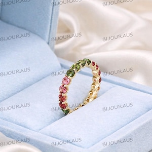 May include: A gold band ring with a continuous row of colorful gemstones. The gemstones are a mix of pink, green, and red. The ring is presented in a light blue jewelry box.
