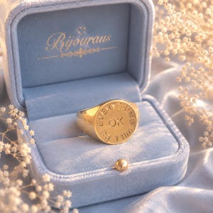 May include: A gold signet ring with the words "EVERYTHING WILL BE OK" engraved on the face. The ring is displayed in a light blue velvet ring box with the brand name "Bijouraus" in gold script.