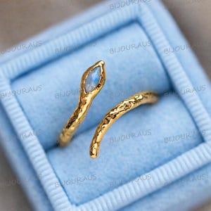May include: A gold-coloured ring in the shape of a snake, with a textured band and a light blue gemstone at the head. The ring is displayed in a light blue velvet box, showcasing its intricate design and craftsmanship.