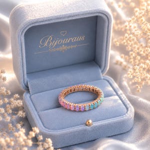 May include: A rose gold eternity ring with a gradient of pink and blue gemstones, presented in a light blue velvet ring box. The box is open, showcasing the ring's colourful stones. The box has the word "Bijouraus" in gold lettering.