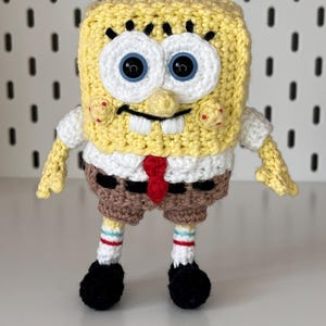May include: A handmade crochet SpongeBob SquarePants figure. The figure is yellow with black eyes, a white shirt, brown shorts, and black shoes. It has a red tie and is standing upright.