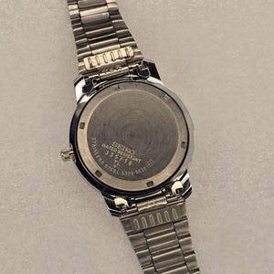 May include: A stainless steel Seiko watch with a silver band. The watch face is round with a black bezel. The back of the watch has the text "SEIKO WATER RESISTANT STAINLESS STEEL 335718 TL 6309-5820".