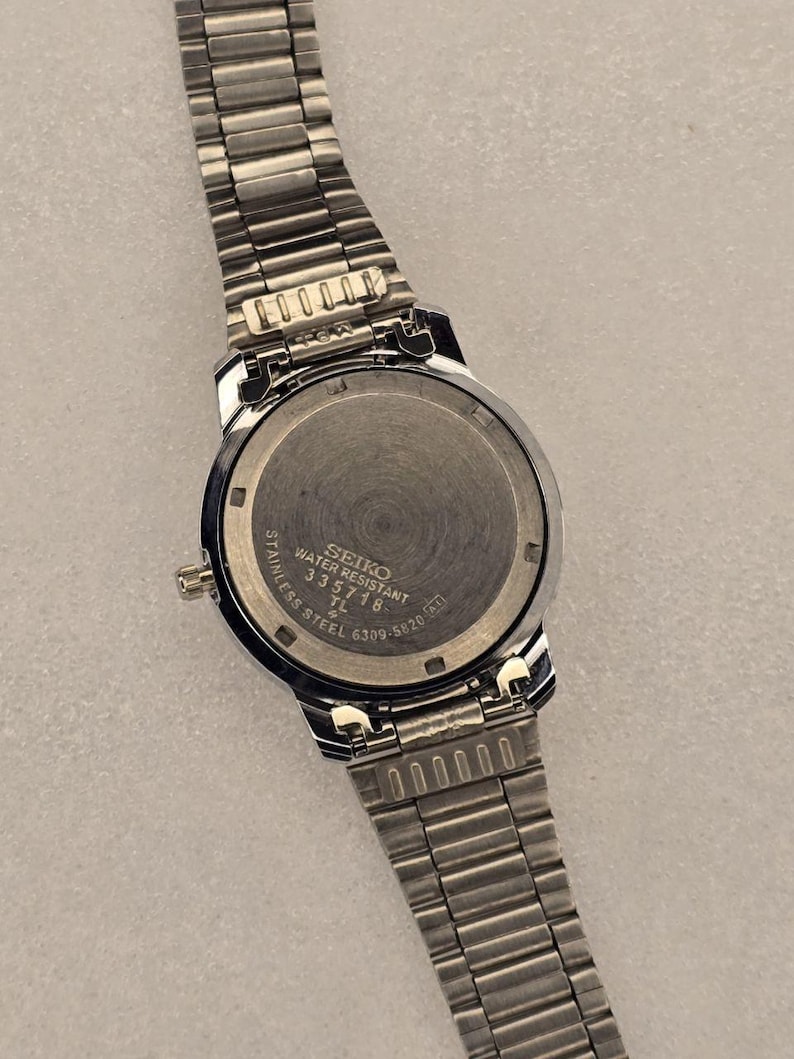 May include: A stainless steel Seiko watch with a silver band. The watch face is round with a black bezel. The back of the watch has the text "SEIKO WATER RESISTANT STAINLESS STEEL 335718 TL 6309-5820".