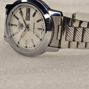 May include: A silver-toned Seiko automatic wristwatch with a white face. The watch features a date display, silver hands, and a silver band. The watch face has black and green markers and the words "Seiko" and "Automatic".