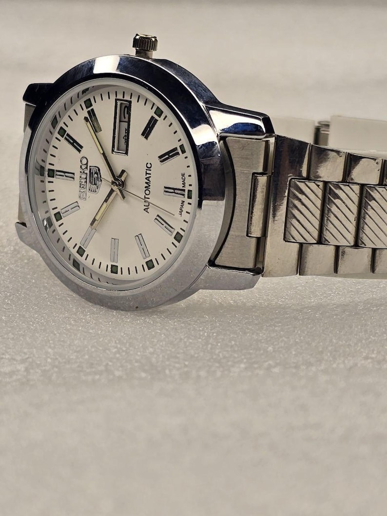 May include: A silver-toned Seiko automatic wristwatch with a white face. The watch features a date display, silver hands, and a silver band. The watch face has black and green markers and the words "Seiko" and "Automatic".