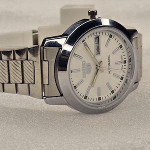 May include: A silver-toned Seiko 5 automatic wristwatch with a white dial. The watch features a date display at the 3 o'clock position and a silver band. The watch is displayed on a light-colored surface.