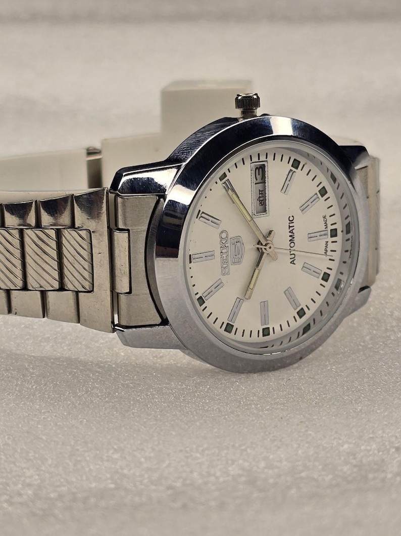May include: A silver-toned Seiko 5 automatic wristwatch with a white dial. The watch features a date display at the 3 o'clock position and a silver band. The watch is displayed on a light-colored surface.