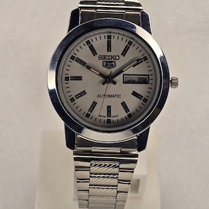 May include: A silver-toned Seiko 5 Automatic wristwatch with a white face. The watch features a day and date display, black hour markers, and a silver band. The watch face displays the text "SEIKO 5 AUTOMATIC JAPAN MADE".