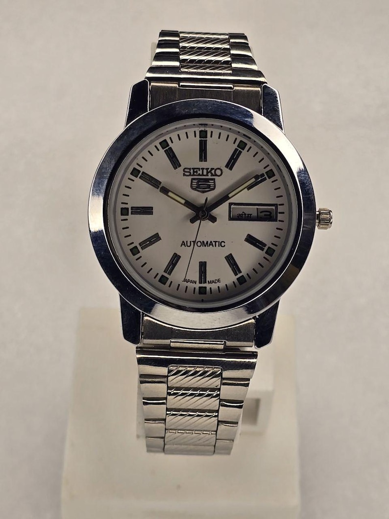 May include: A silver-toned Seiko 5 Automatic wristwatch with a white face. The watch features a day and date display, black hour markers, and a silver band. The watch face displays the text "SEIKO 5 AUTOMATIC JAPAN MADE".