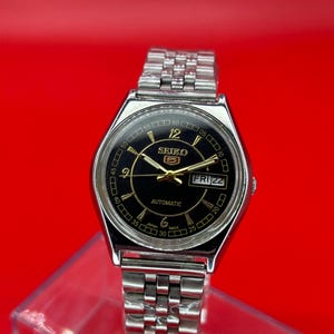 May include: A silver-toned Seiko 5 automatic wristwatch with a black dial. The watch features gold-colored hands and markers, a day-date display, and a stainless steel band. The watch face displays the text "SEIKO 5" and "AUTOMATIC".