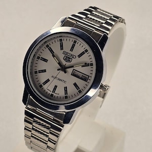 May include: A silver Seiko 5 automatic wristwatch with a light-colored face. The watch has a stainless steel band and a date display. The face features black markers and hands, with the Seiko logo prominently displayed.