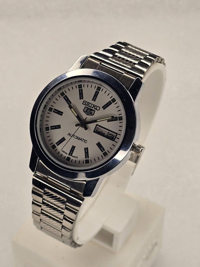 May include: A silver Seiko 5 automatic wristwatch with a light-colored face. The watch has a stainless steel band and a date display. The face features black markers and hands, with the Seiko logo prominently displayed.