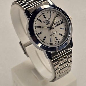 May include: A silver-toned Seiko 5 automatic wristwatch. The watch has a white face with black hour markers and hands, and a day-date display. The watchband is made of silver-toned metal links. The watch is displayed on a white stand.