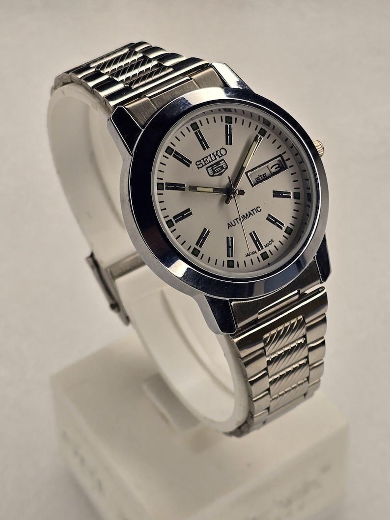 May include: A silver-toned Seiko 5 automatic wristwatch. The watch has a white face with black hour markers and hands, and a day-date display. The watchband is made of silver-toned metal links. The watch is displayed on a white stand.