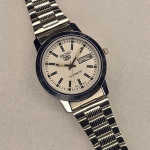 May include: A silver-toned Seiko 5 Automatic wristwatch with a white face, black markers, and a day-date display. The watch has a stainless steel band with a textured design. The watch face reads "SEIKO 5 AUTOMATIC" and "JAPAN MADE".