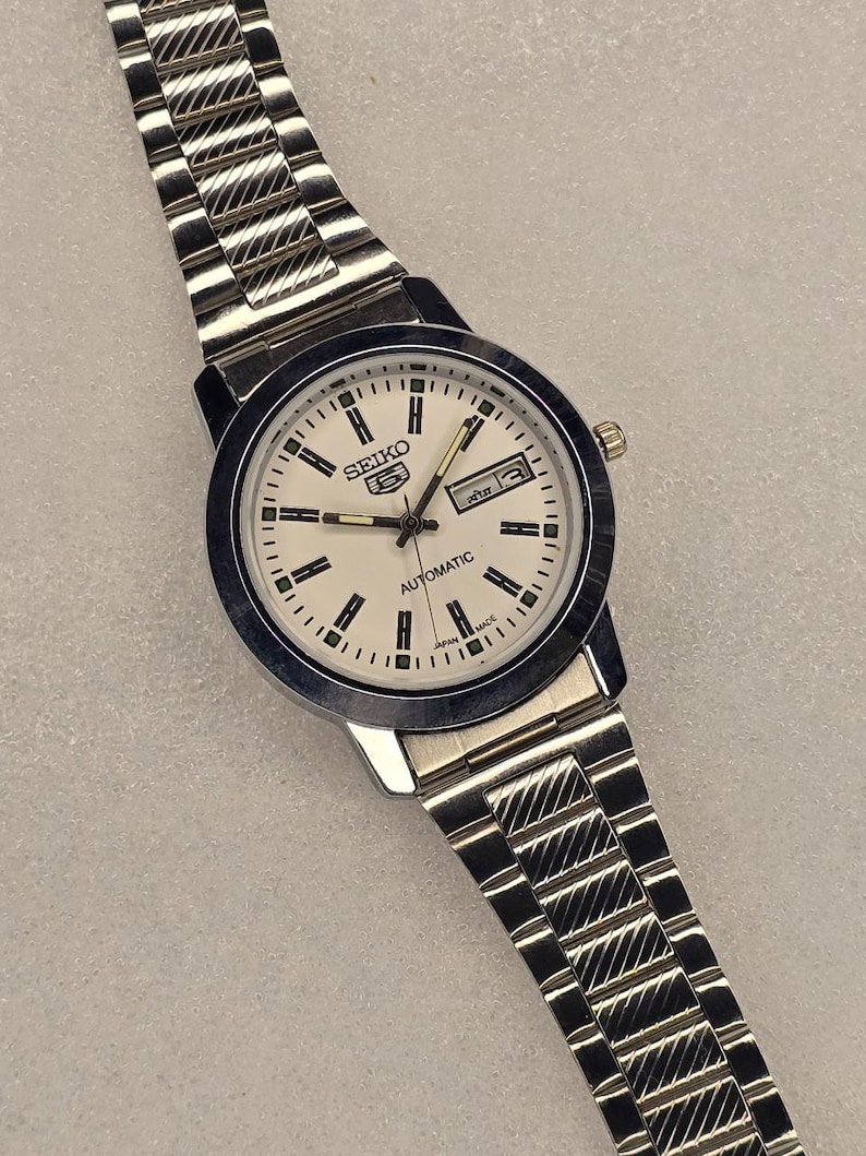 May include: A silver-toned Seiko 5 Automatic wristwatch with a white face, black markers, and a day-date display. The watch has a stainless steel band with a textured design. The watch face reads "SEIKO 5 AUTOMATIC" and "JAPAN MADE".