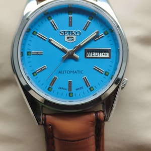 May include: A silver Seiko 5 automatic watch with a vibrant blue face and brown leather strap. The watch face shows the day and date, with silver hands and markers. The watch is marked "Japan Made".