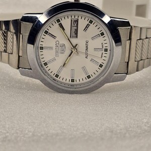 May include: A silver-toned Seiko 5 automatic wristwatch with a white face. The watch features a day and date display, luminous hands, and a stainless steel band. The watch is a classic design.