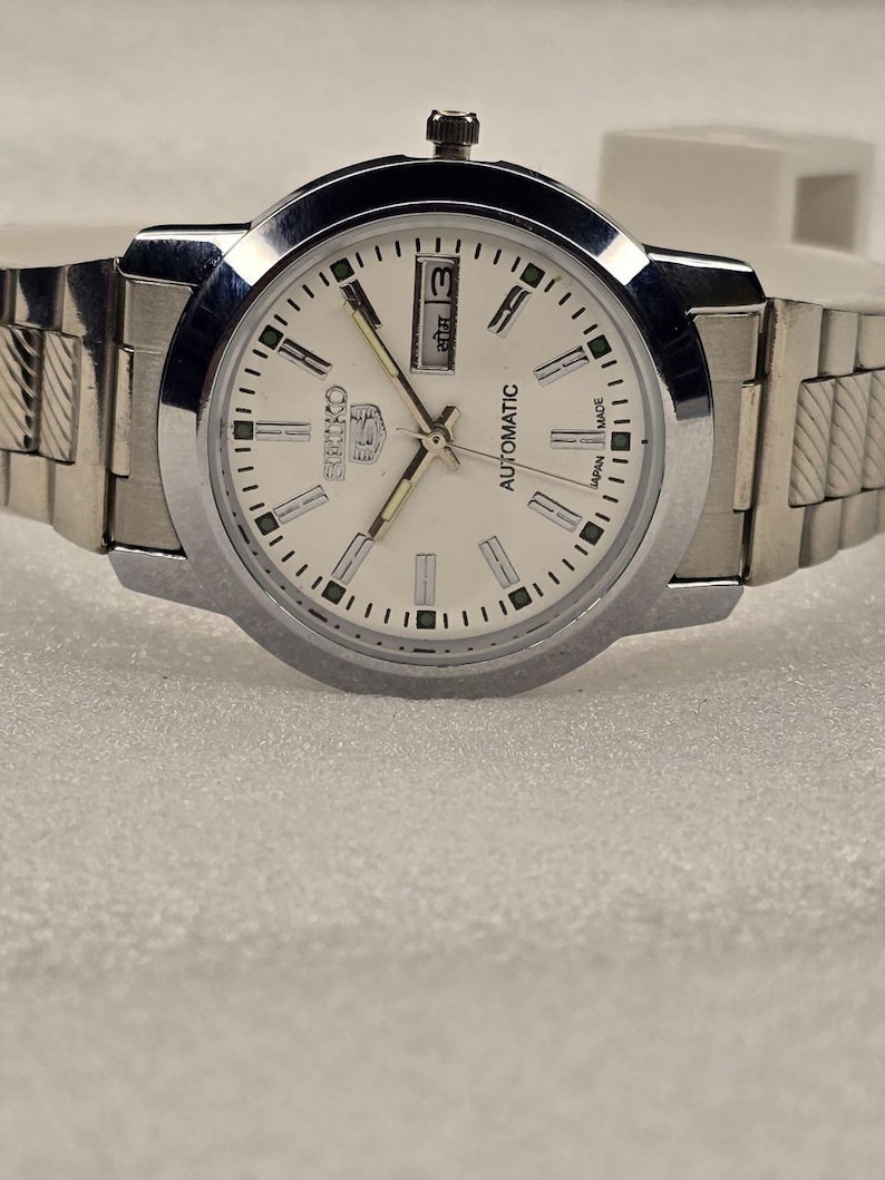 May include: A silver-toned Seiko 5 automatic wristwatch with a white face. The watch features a day and date display, luminous hands, and a stainless steel band. The watch is a classic design.