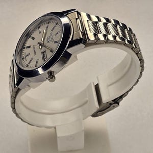 May include: A silver-toned wristwatch with a light-colored face and a metal band. The watch features a day and date display and is labeled "Automatic". The watch is displayed on a white stand.