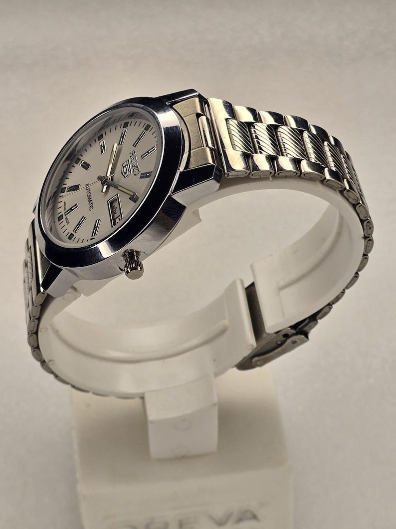 May include: A silver-toned wristwatch with a light-colored face and a metal band. The watch features a day and date display and is labeled "Automatic". The watch is displayed on a white stand.
