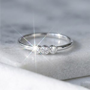 Peut inclure: Une bague en argent ornée de trois pierres précieuses rondes et transparentes. La bague est posée sur une surface blanche et scintille à la lumière. Les pierres sont espacées uniformément, pour un look classique et élégant.