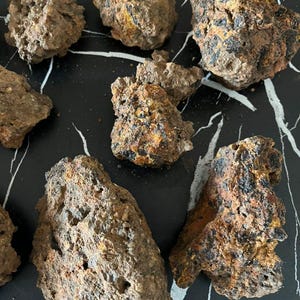 Pode incluir: Uma coleção de sete rochas irregulares e porosas com uma textura áspera e escarpada. As pedras exibem uma paleta de cores manchadas de cinza carvão escuro, marrom terroso e manchas de laranja ferrugem oxidado com cavidades.