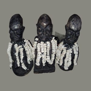 May include: Three dark wooden sculptures of human figures. Each figure has a carved face and is adorned with strands of white shell beads around the neck and chest. The sculptures are displayed against a neutral background.