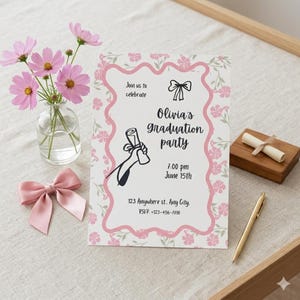 May include: A graduation party invitation with a pink floral border and the text "Olivia's Graduation Party." The invitation includes the date, time, and RSVP information. A pink bow, flowers in a vase, a pen, and a small wooden box are also present.