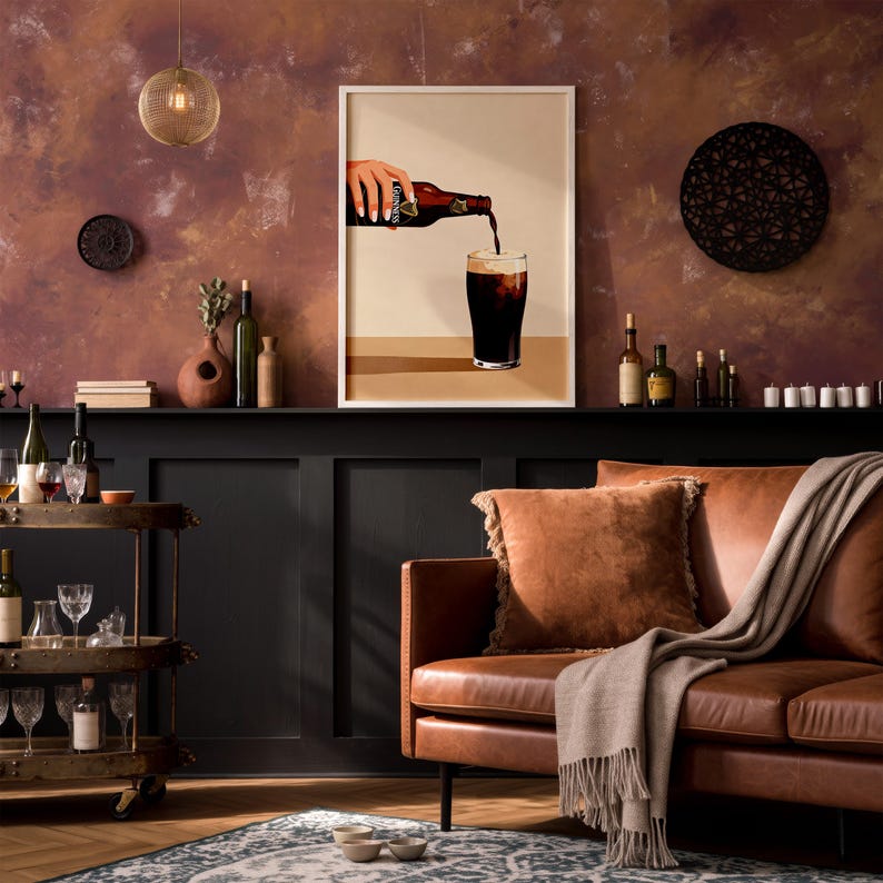 May include: A framed print depicts a hand pouring dark beer into a glass. The image is set against a warm, textured wall. The room features a brown leather sofa, a bar cart with glassware, and decorative items.