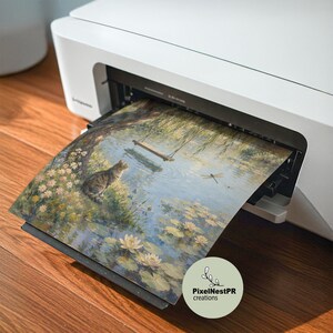 May include: A white inkjet printer outputting a printed sheet featuring a painting of a tabby cat sitting by a pond with water lilies, a hanging rope swing, and a dragonfly. A circular logo with the text PixelNestPR creations is visible.