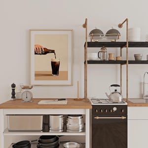 May include: A kitchen scene with a framed print of a hand pouring a dark beverage into a glass. The kitchen features a wooden countertop, a stove, and open shelving with kitchenware. A scale and other kitchen tools are also visible.
