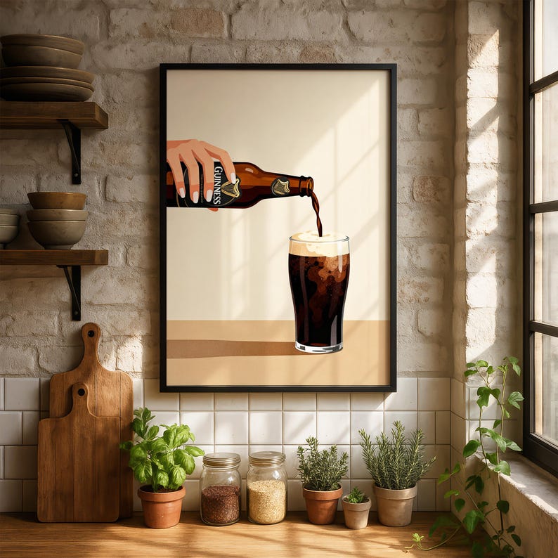 May include: Framed art print featuring a hand pouring a dark stout from a Guinness bottle into a glass. The beer has a creamy, white head. The artwork is set against a neutral background with a black frame, suitable for a kitchen or bar area.