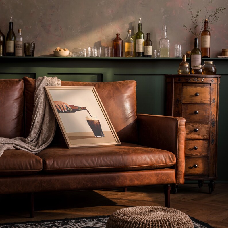 May include: A framed art print depicting a hand pouring a dark beverage into a glass, resting on a brown leather sofa. The room features a wooden cabinet, bottles, and glasses on a shelf, creating a cozy, homey atmosphere.