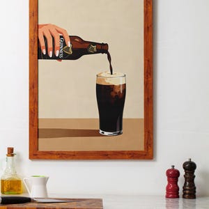 May include: A framed print depicts a hand pouring Guinness stout from a bottle into a glass. The bottle is brown with the Guinness label. The glass is filled with dark liquid and a creamy head. The frame is a warm brown color.