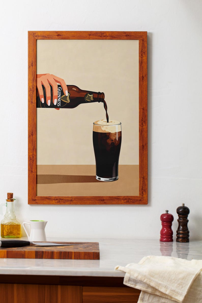 May include: A framed print depicts a hand pouring Guinness stout from a bottle into a glass. The bottle is brown with the Guinness label. The glass is filled with dark liquid and a creamy head. The frame is a warm brown color.
