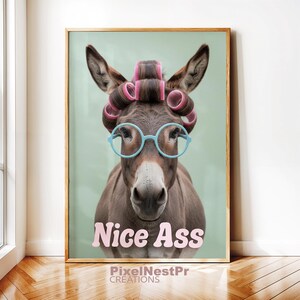 May include: A framed print featuring a donkey wearing pink hair rollers and light blue glasses. The donkey has the words "Nice Ass" printed in pink. The background is a light green color.