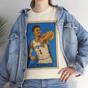 May include: A cream-coloured t-shirt with a basketball player illustration in a blue frame. The player wears a Spurs jersey with the number 1. The text "EL CODAZO" is at the bottom. The shirt is worn with a light blue denim jacket.