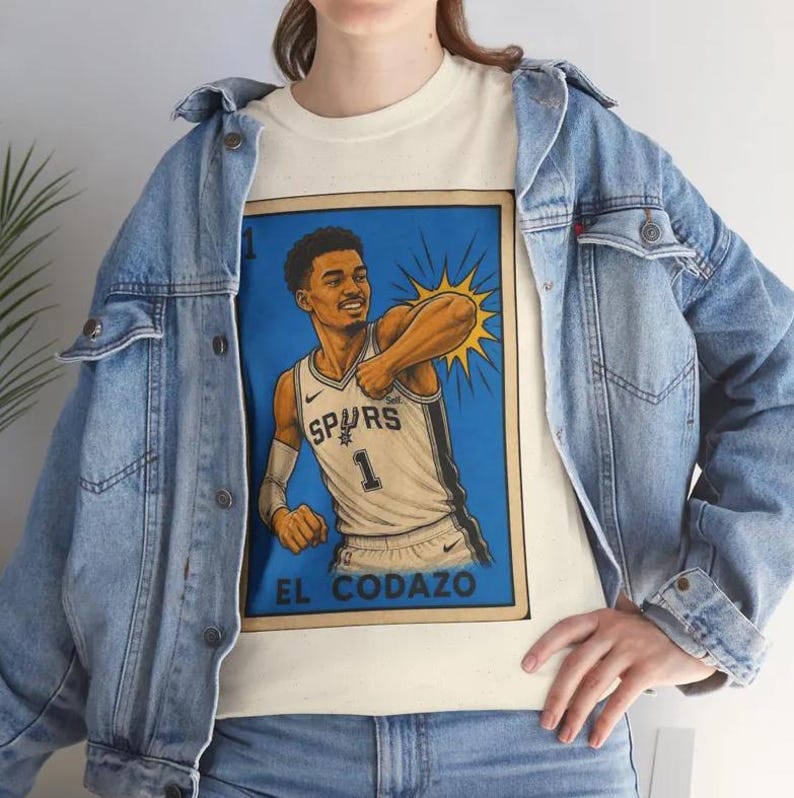 May include: A cream-coloured t-shirt with a basketball player illustration in a blue frame. The player wears a Spurs jersey with the number 1. The text "EL CODAZO" is at the bottom. The shirt is worn with a light blue denim jacket.