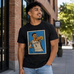 May include: Black t-shirt with a graphic of a basketball player in a Spurs jersey. The graphic is in a blue frame with the text "EL CODAZO". The player is shown with a burst of yellow light.