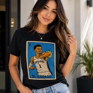 May include: Black t-shirt with a graphic of a basketball player in a Spurs jersey, with the text "EL CODAZO" below. The graphic is framed in a blue border. The shirt is worn by a person in blue jeans.