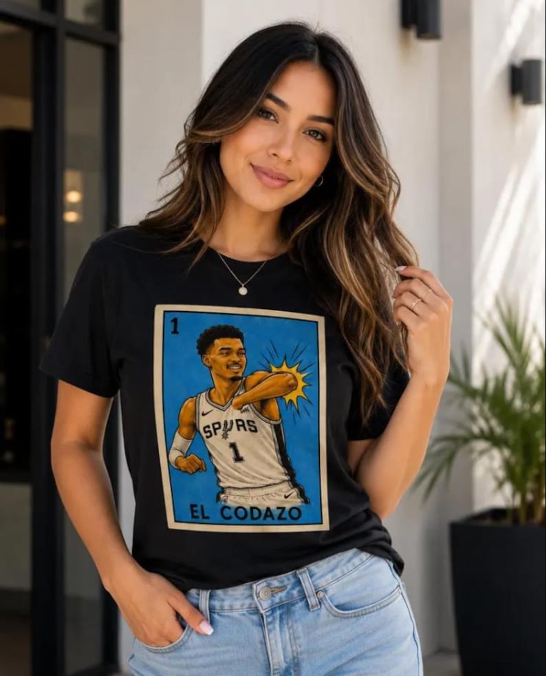 May include: Black t-shirt with a graphic of a basketball player in a Spurs jersey, with the text "EL CODAZO" below. The graphic is framed in a blue border. The shirt is worn by a person in blue jeans.