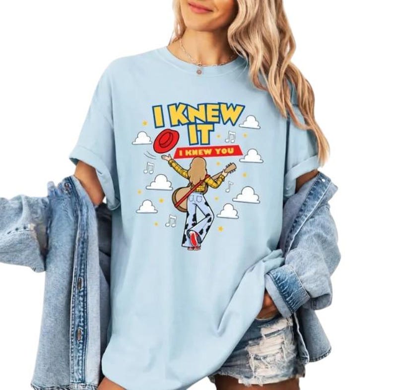 Pu&ograve; includere: Una t-shirt azzurro chiaro con la grafica di una persona dai capelli lunghi che suona la chitarra, indossando una camicia a quadri gialla e pantaloni con stampa muccata. Il testo recita I KNEW IT e I KNEW YOU, tra nuvole bianche e note.