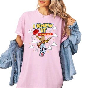 Pu&ograve; includere: Una t-shirt rosa chiaro con la grafica di una persona che suona una chitarra acustica, indossando una camicia a quadri gialla e pantaloni con stampa muccata. Il testo recita I KNEW IT e I KNEW YOU, circondato da nuvole e note musicali.