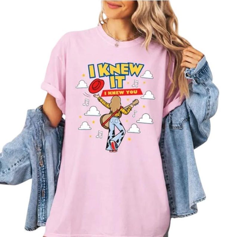 Pu&ograve; includere: Una t-shirt rosa chiaro con la grafica di una persona che suona una chitarra acustica, indossando una camicia a quadri gialla e pantaloni con stampa muccata. Il testo recita I KNEW IT e I KNEW YOU, circondato da nuvole e note musicali.